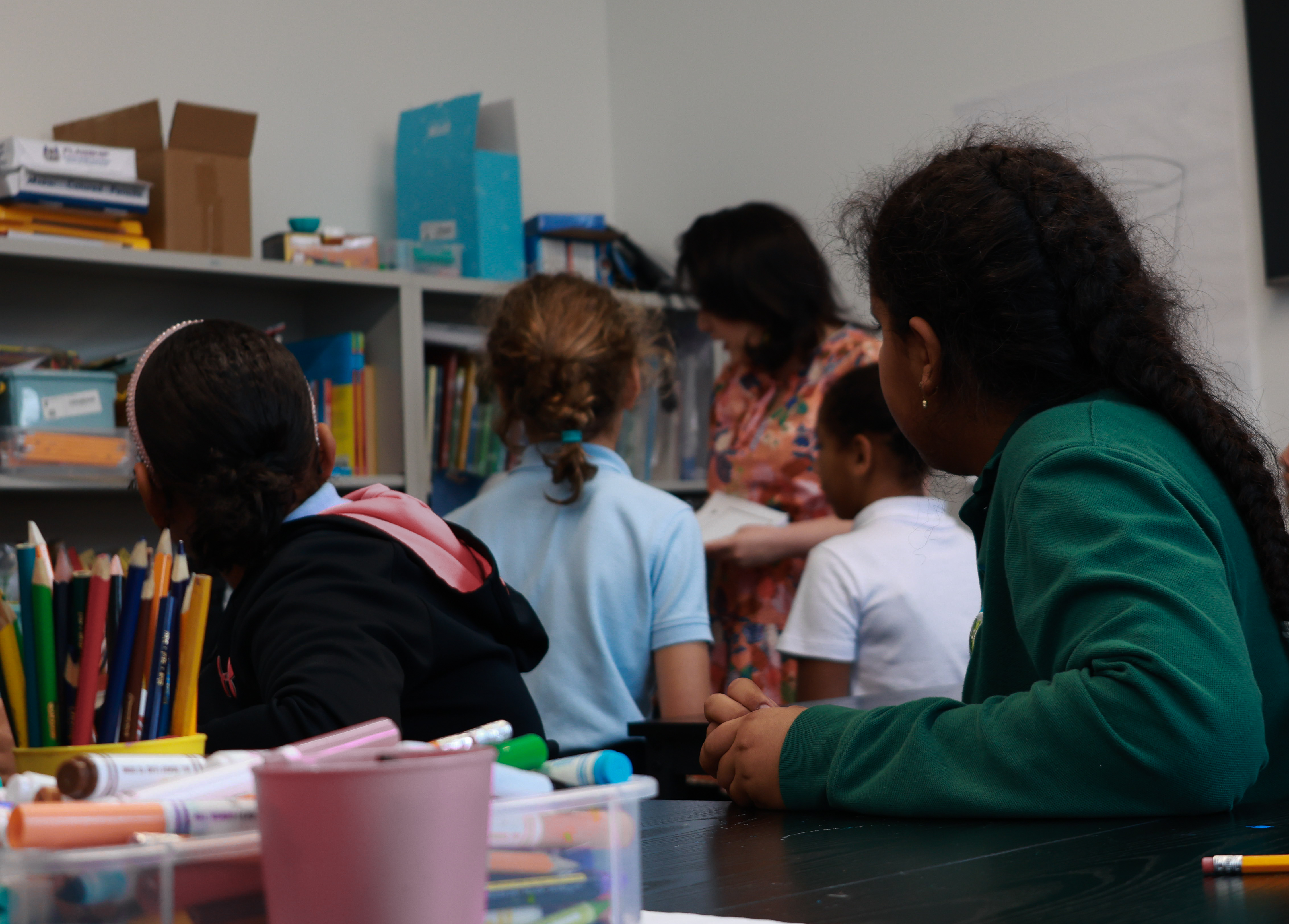 A group of students sitting at tables with art supplies face away from the camera toward a teacher standing in a coral-colored dress.