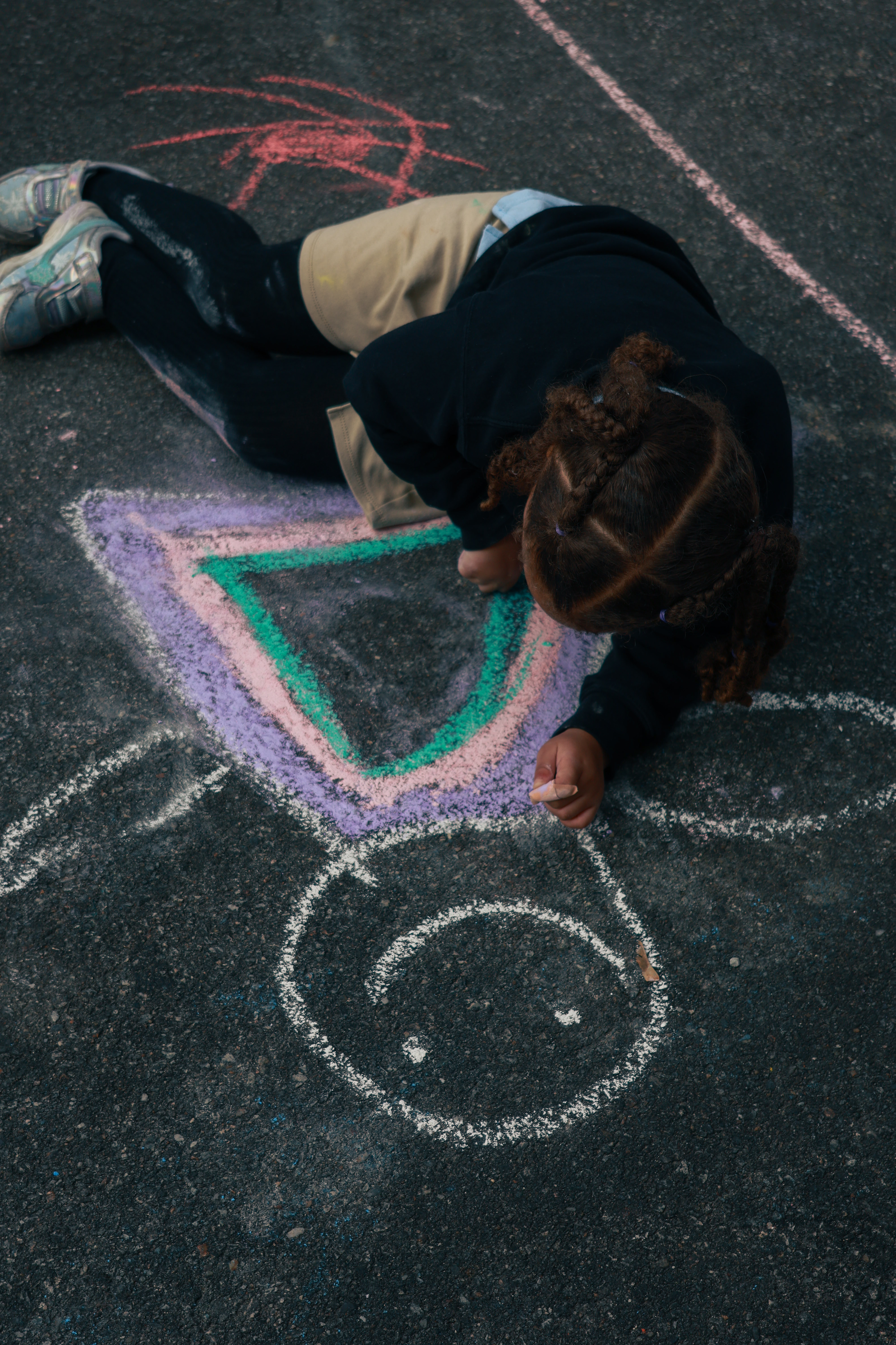 A child in a school uniform lays on the pavement using chalk to draw a fairy-like figure in a pink, purple and green dress.