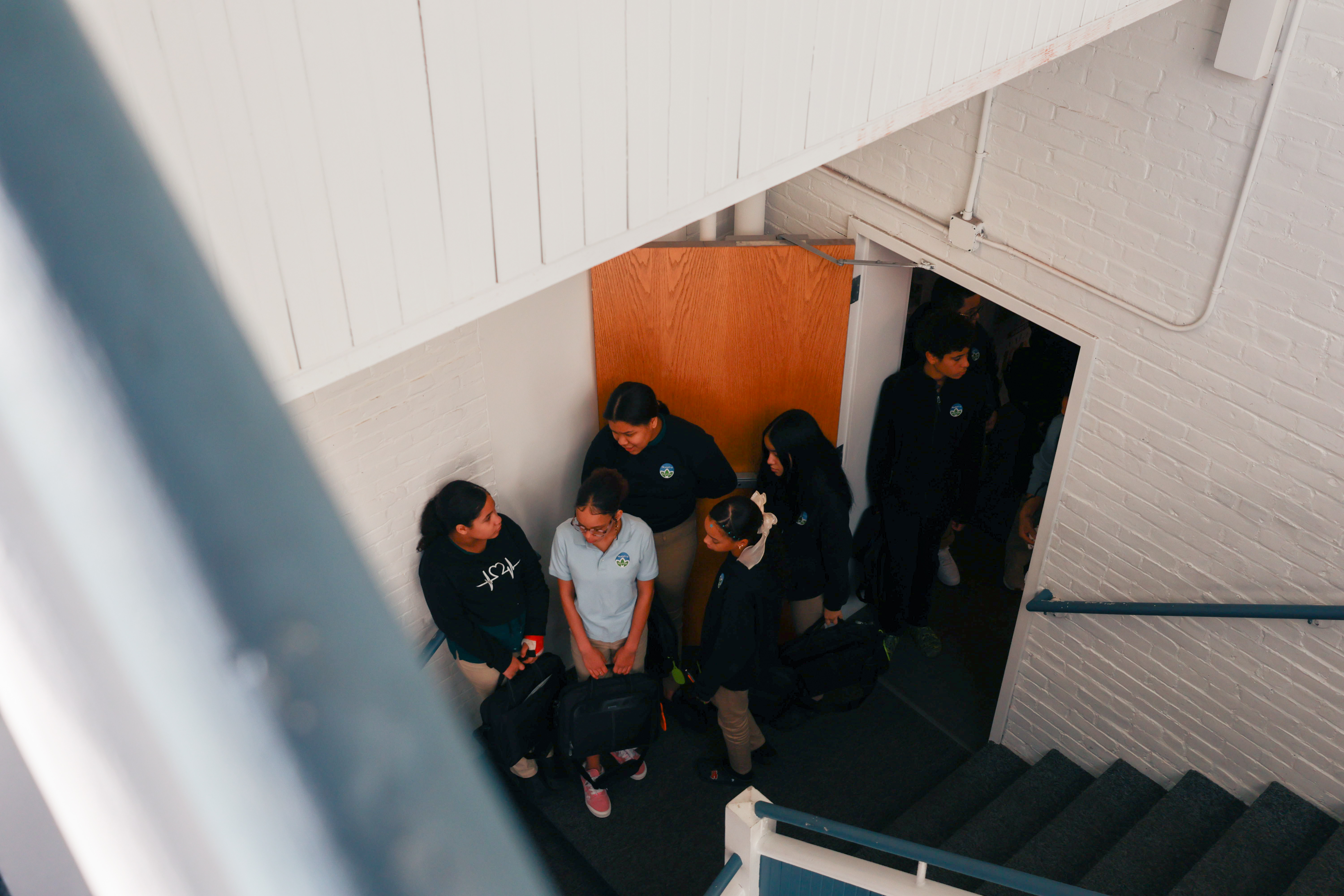 Photo of students waiting and talking to each other at the base of a stairwell wearing their uniforms.