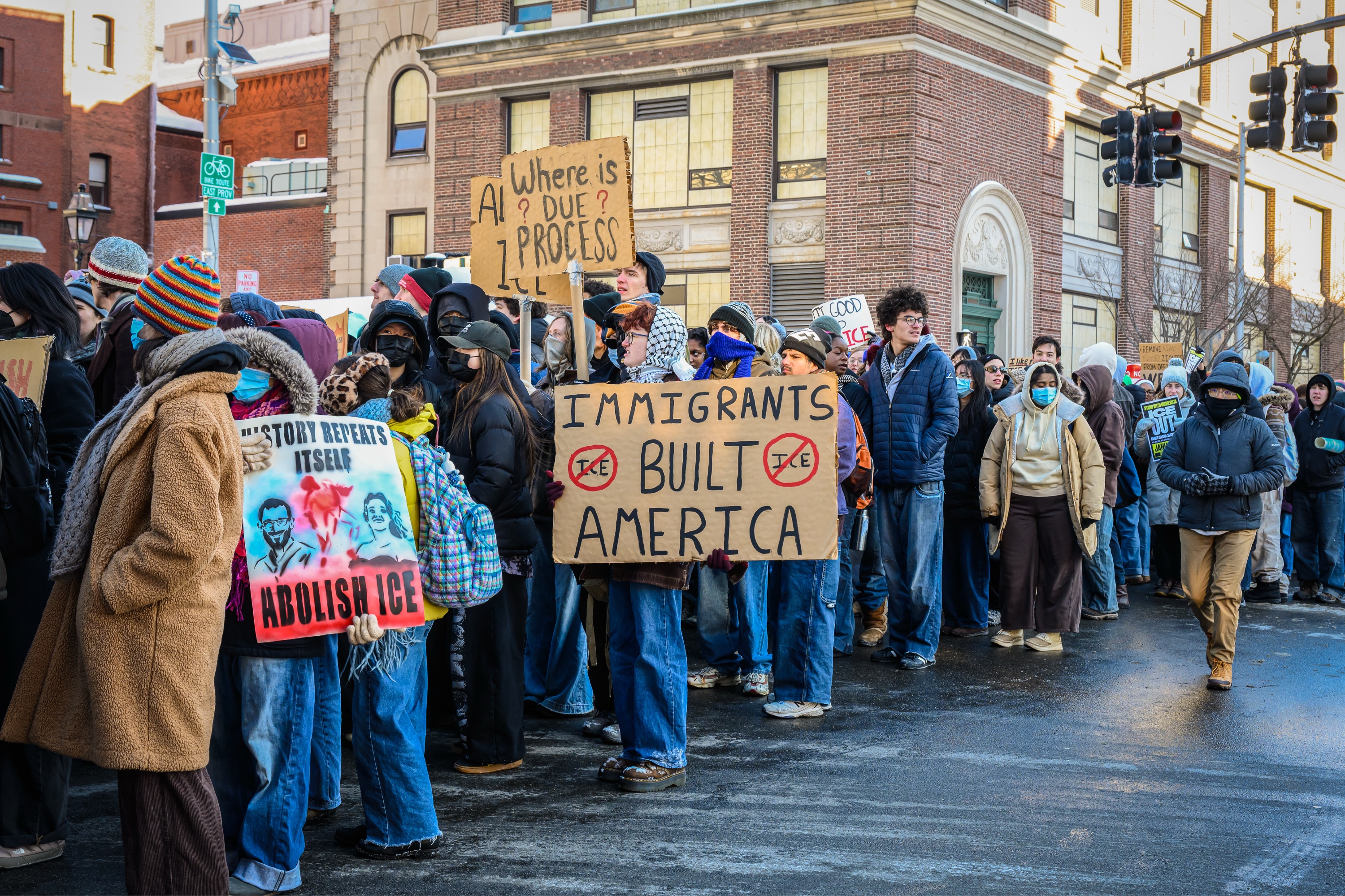 A crowd of protesters walking past a street holding posters stating “HISTORY REPEATS ITSELF / ABOLISH ICE,” “IMMIGRANTS / BUILT / AMERICA,” and “WHERE IS / ? DUE ? / PROCESS”