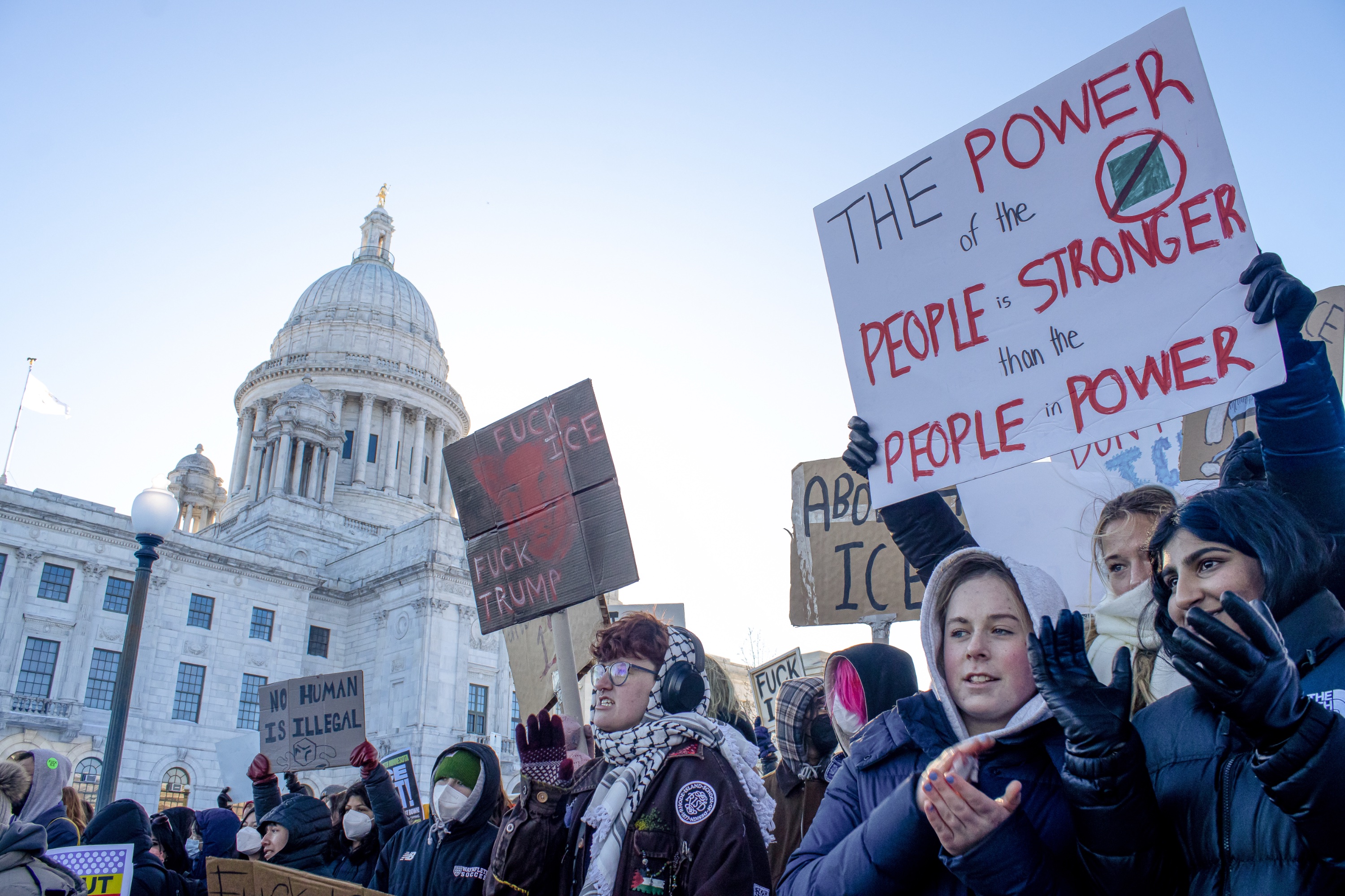 A group of protesters stand outside of the Rhode Island State House One protester holds up a sign that reads, “The power of the people is stronger than the people in power.”