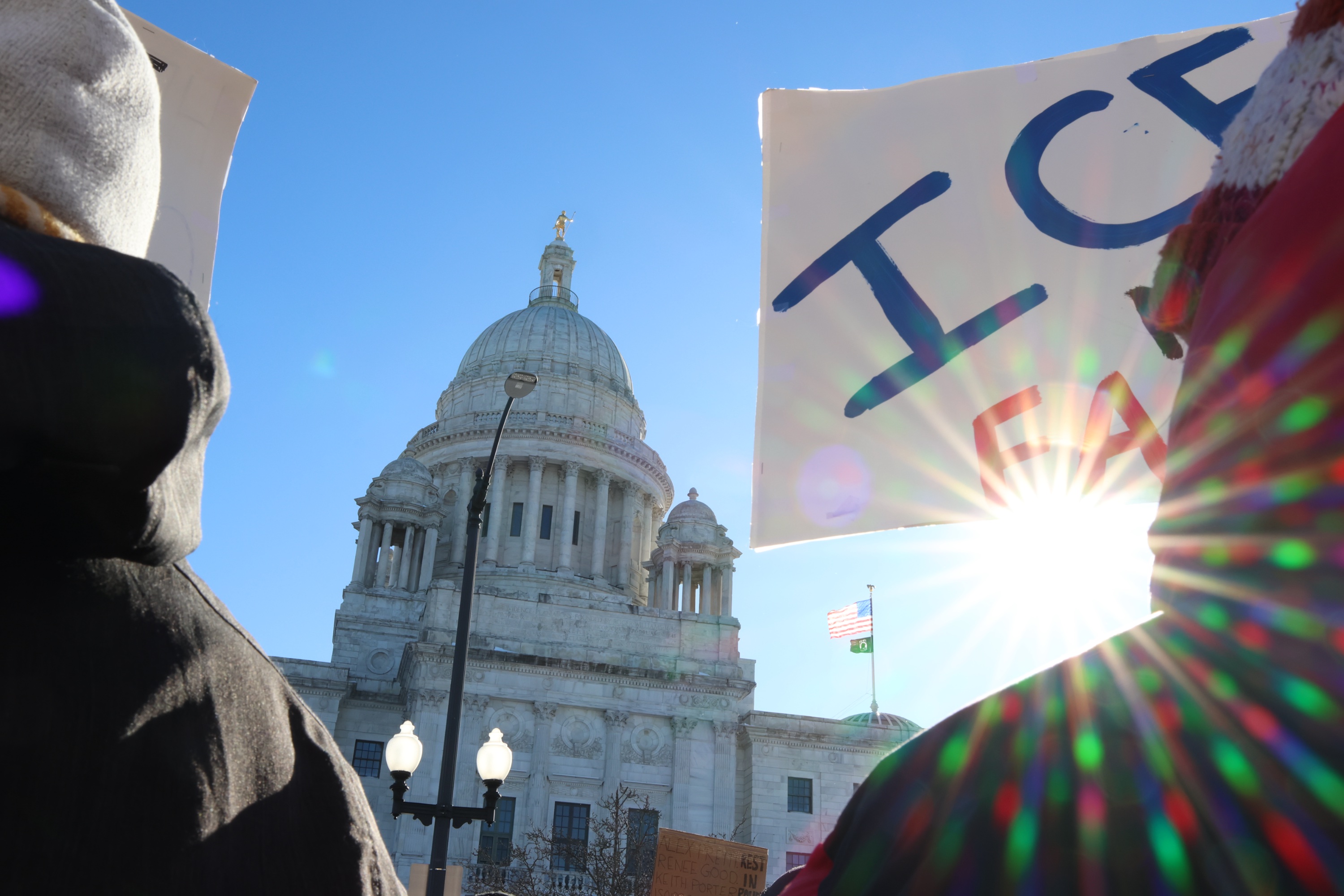 Protesters hold up signs at the State House. The sun shines behind the State House.