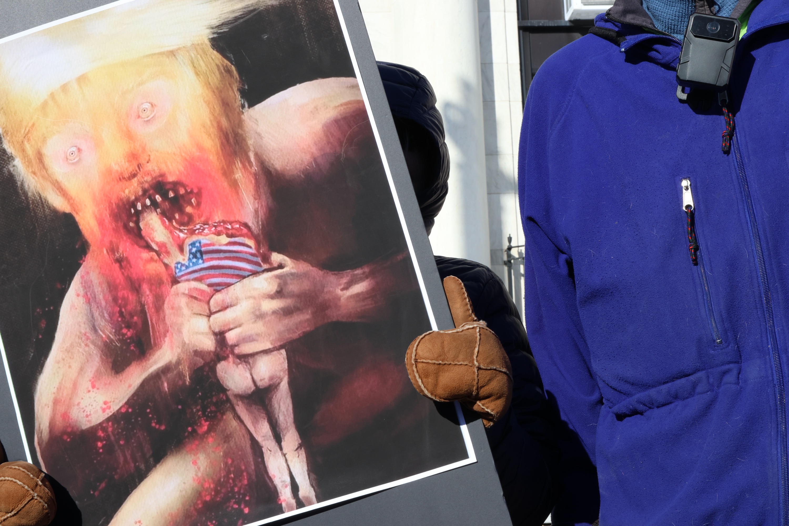 A protester holding a sign with a painting of a big, blonde man eating a human with the American flag on its back. The painting resembles Goya’s “Saturn Devouring His Son”