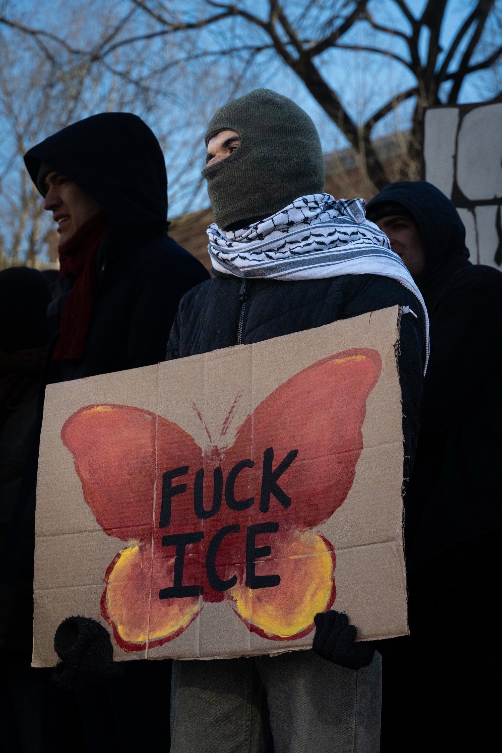 A masked protester holds up a poster that reads, “Fuck ICE” inside of a red painted butterfly.