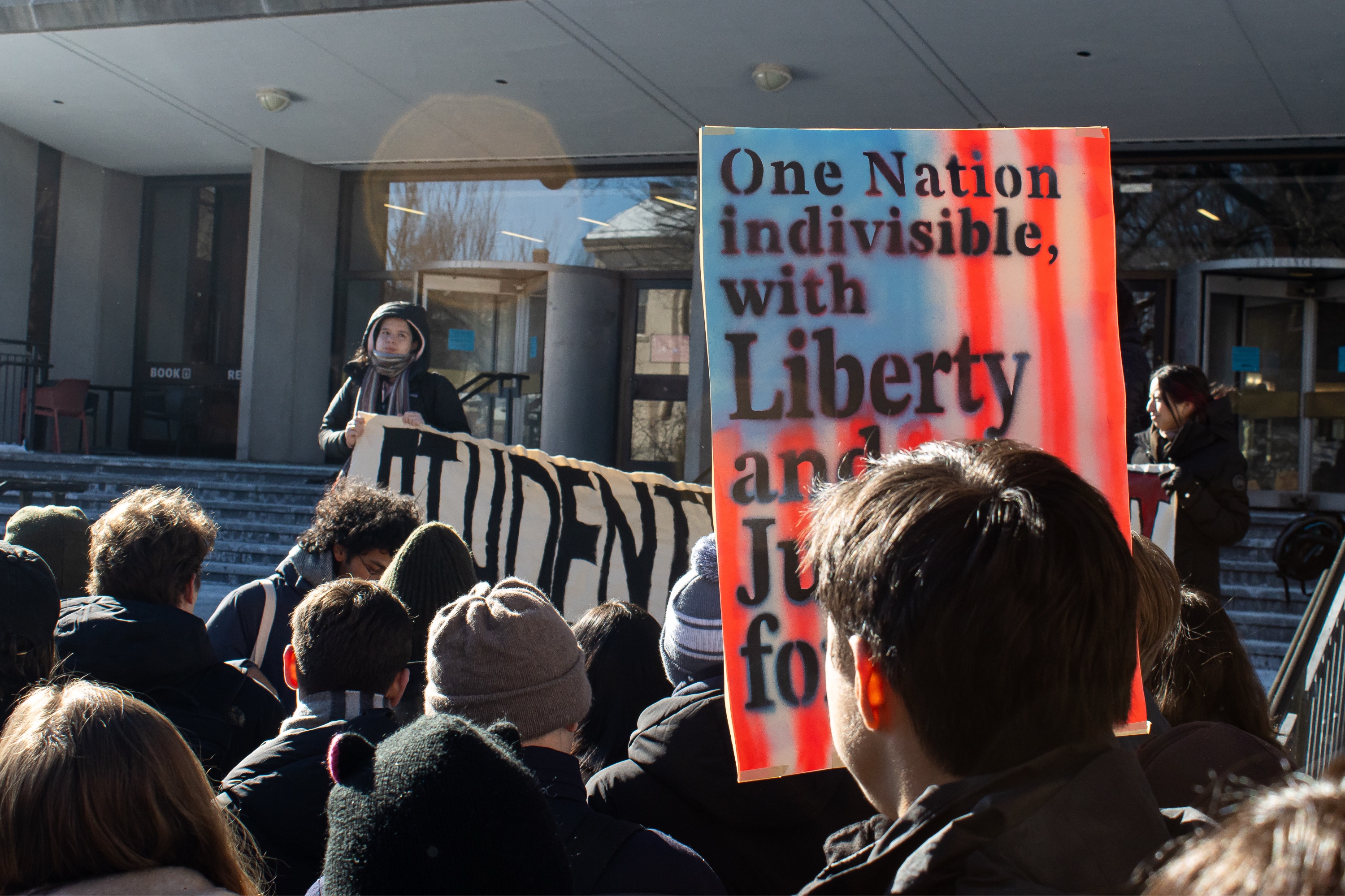 A group of protestors stand outside the John D. Rockefeller Library upholding a sign that reads, “One nation indivisible, with liberty and justice for all.”